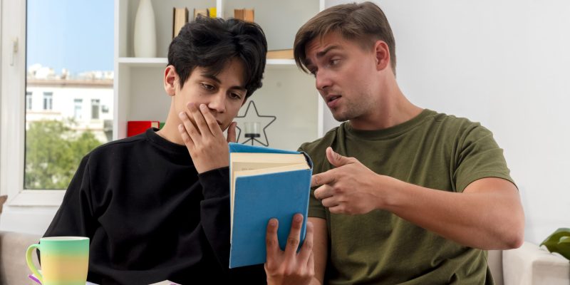 confident young blonde handsome man holding and pointing at book sitting at table and looking at young brunette handsome guy putting hand on mouth looking at book inside design living room