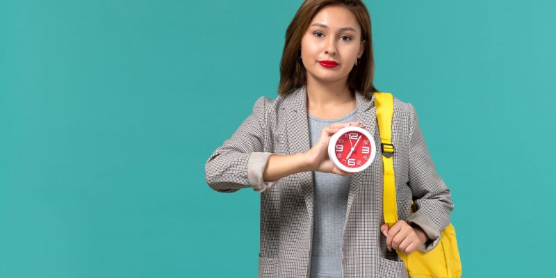 front-view-female-student-grey-jacket-wearing-her-yellow-backpack-holding-clocks-light-blue-wall